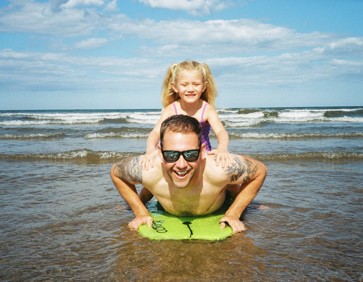 A dad uses a body board in the sea with his young daughter.